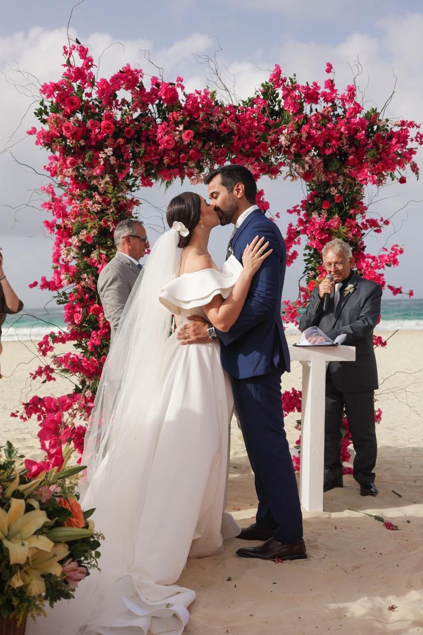 Noivos se beijam sob um arco de flores em uma cerimônia na praia